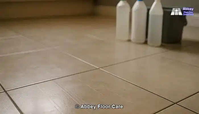 Close-up of a tiled floor with dull traffic lanes and hazy residue, with generic cleaning products blurred in the background.